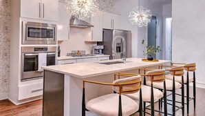 A modern kitchen with stainless steel appliances, a central island with bar stools, and decorative crystal chandeliers. - Gulch Oasis • 15 Beds • Rooftop Lounge w/ Fire Pit (Nashville)
