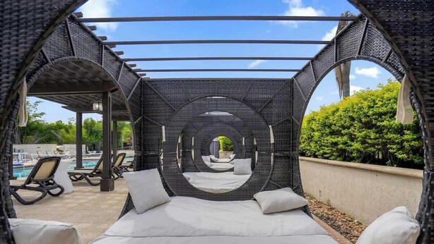 Cama com lençóis brancos em estrutura circular de vime, com vista para uma piscina e espreguiçadeiras.