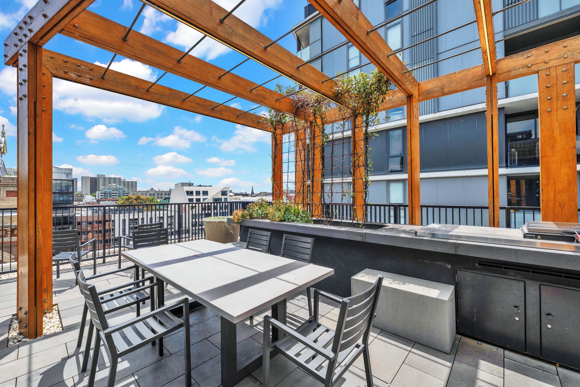 An outdoor terrace with a table and chairs, a wooden pergola, and a view of a cityscape.