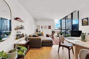A modern living room with a sofa, a dining area with a table and chairs, a large window with a city view, and a desk with a computer. - Stylish Inner City Apartment (Collingwood)