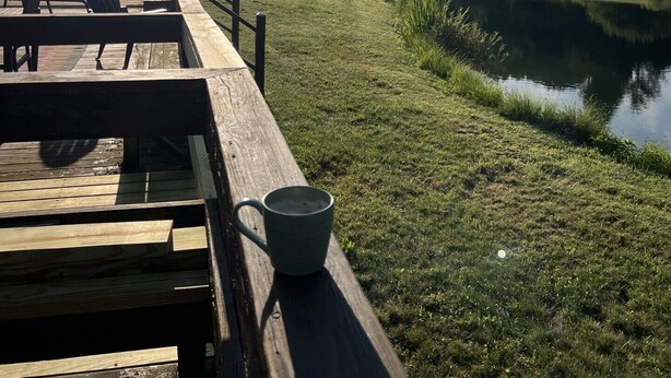 A wooden deck with picnic tables and chairs, overlooking a calm pond and grassy area with trees in the background. - On Wyman Pond. Home on sprawling property with a pond, hiking & wildlife. (Sidney Center)