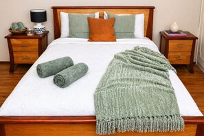Een netjes opgemaakt bed met een witte deken, twee opgerolde handdoeken en een groene plaid.