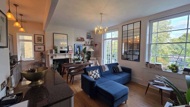Ein geräumiges Wohnzimmer mit einem blauen Sofa, einem Essbereich und einer durch ein großes Fenster sichtbaren Küche.