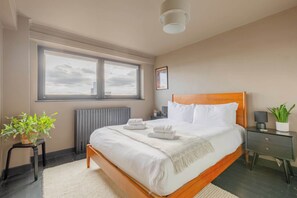 A bedroom with a large window, a bed with white linens, a bedside table with a lamp, and a radiator. - Gorgeously Designed 2BD Flat, Heart of East London (Greater London)