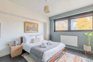 A modern bedroom with a bed, bedside table, lamp, radiator, window, and a framed picture on the wall.