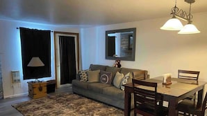 A living room with a sofa, a dining table with chairs, a rug, a lamp, and a mirror on the wall.