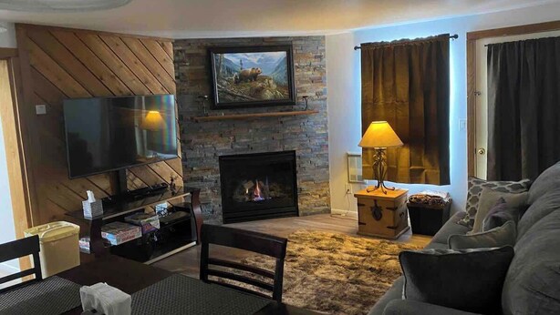 A living room with a fireplace, a television mounted on a wooden wall, a sofa, and a table with a lamp.