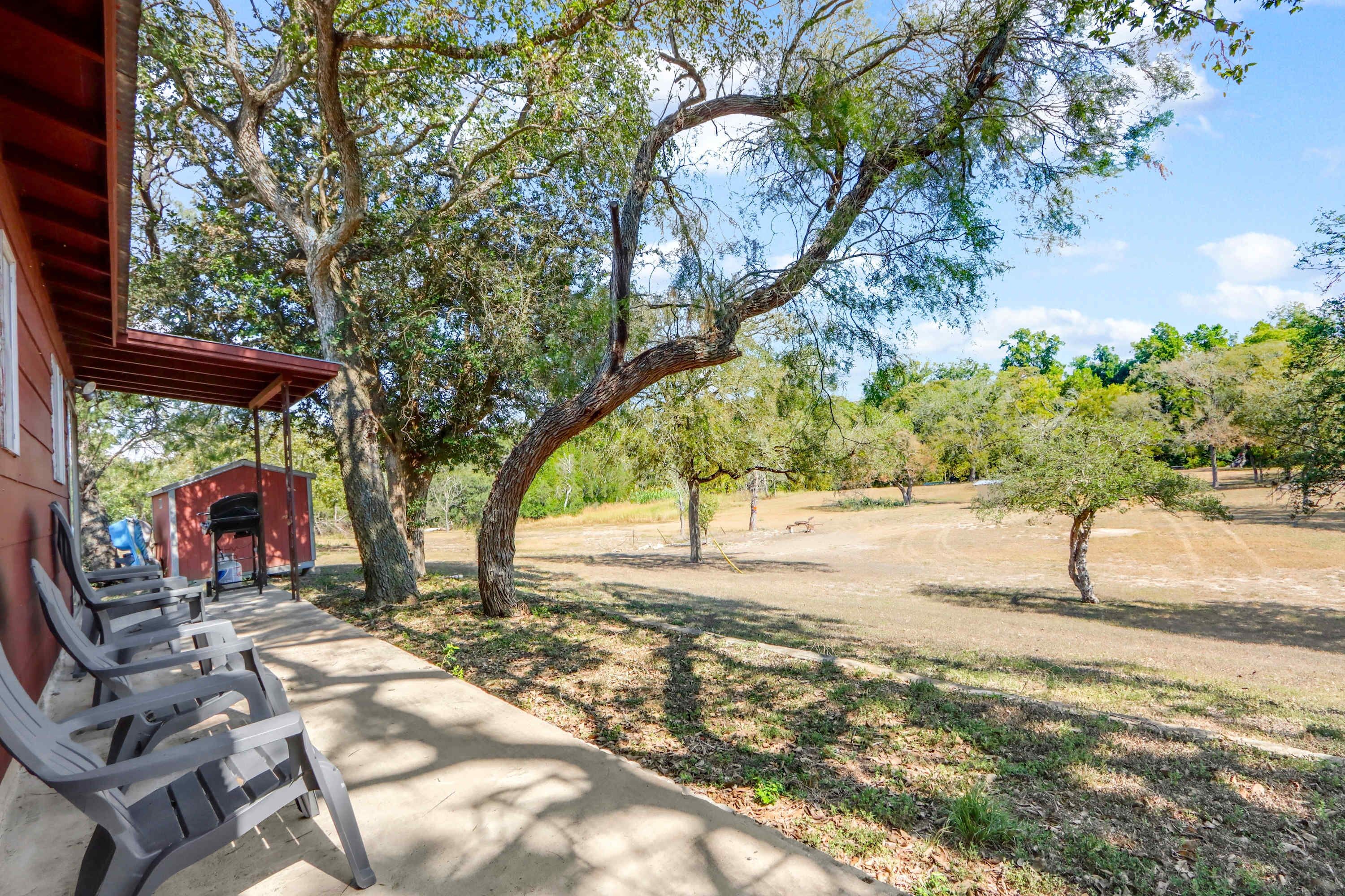 A covered outdoor seating area with chairs, a red building, and trees in a sunny landscape.