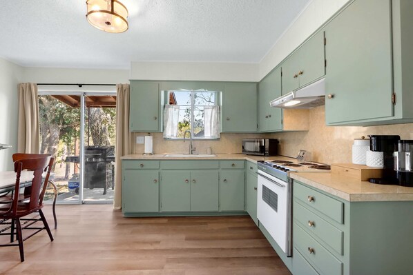 A kitchen with light green cabinets, a large window, and a dining area with wooden chairs.