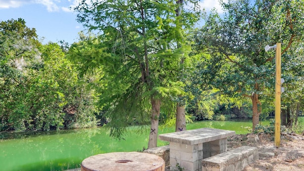 A rustic outdoor seating area with a concrete table and bench, surrounded by trees and a body of water. - Guadalupe Bend Hideaway For 5 (New Braunfels)