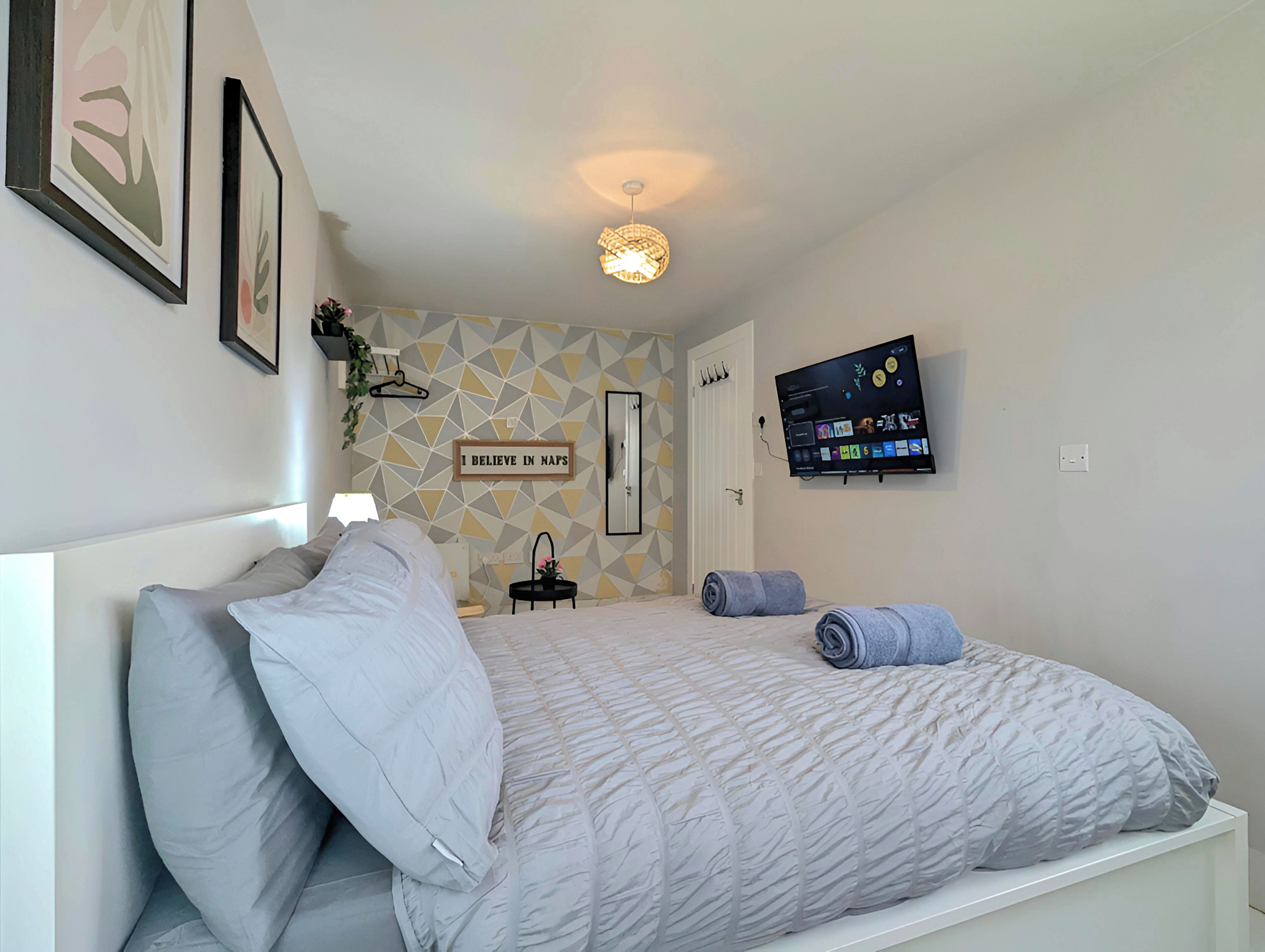 A bedroom with a bed, a wall-mounted TV, a geometric-patterned wall, and a sign that reads 'I BELIEVE IN MAPS'.