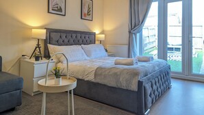 A bedroom with a large bed, bedside tables, a small round table with a plant, and a view of the outdoors through a sliding glass door. - This is a large three-story house, located near the city centre. A short 4-minute drive (1 mile) will take you to the city centre, Albert Dock, ACC. Convenient public transportation is available with a 10-minute bus ride from the city. centre. (Merseyside)