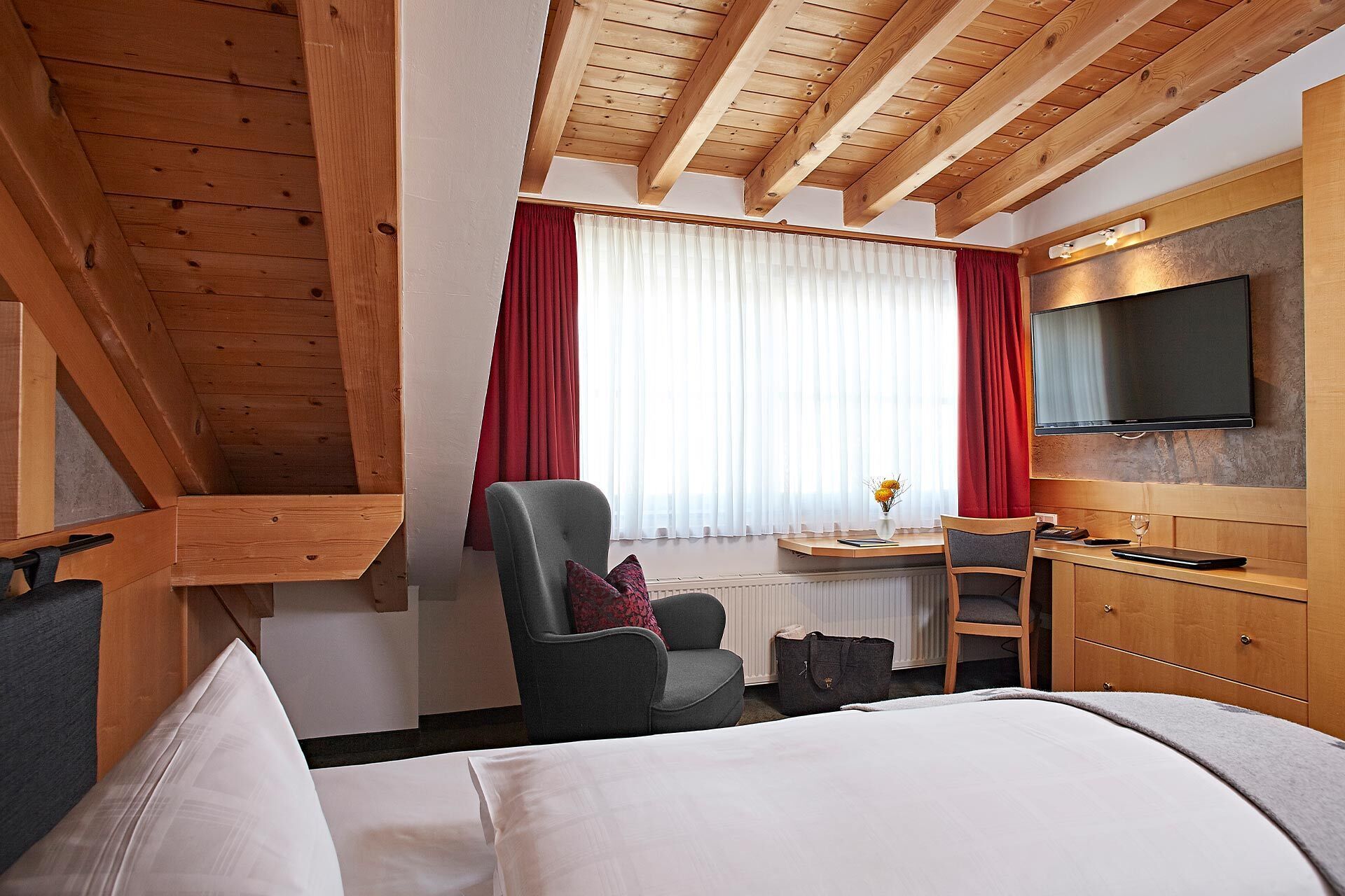 Quarto de hotel com cama, escrivaninha com cadeira, televisão na parede e janela com cortinas.