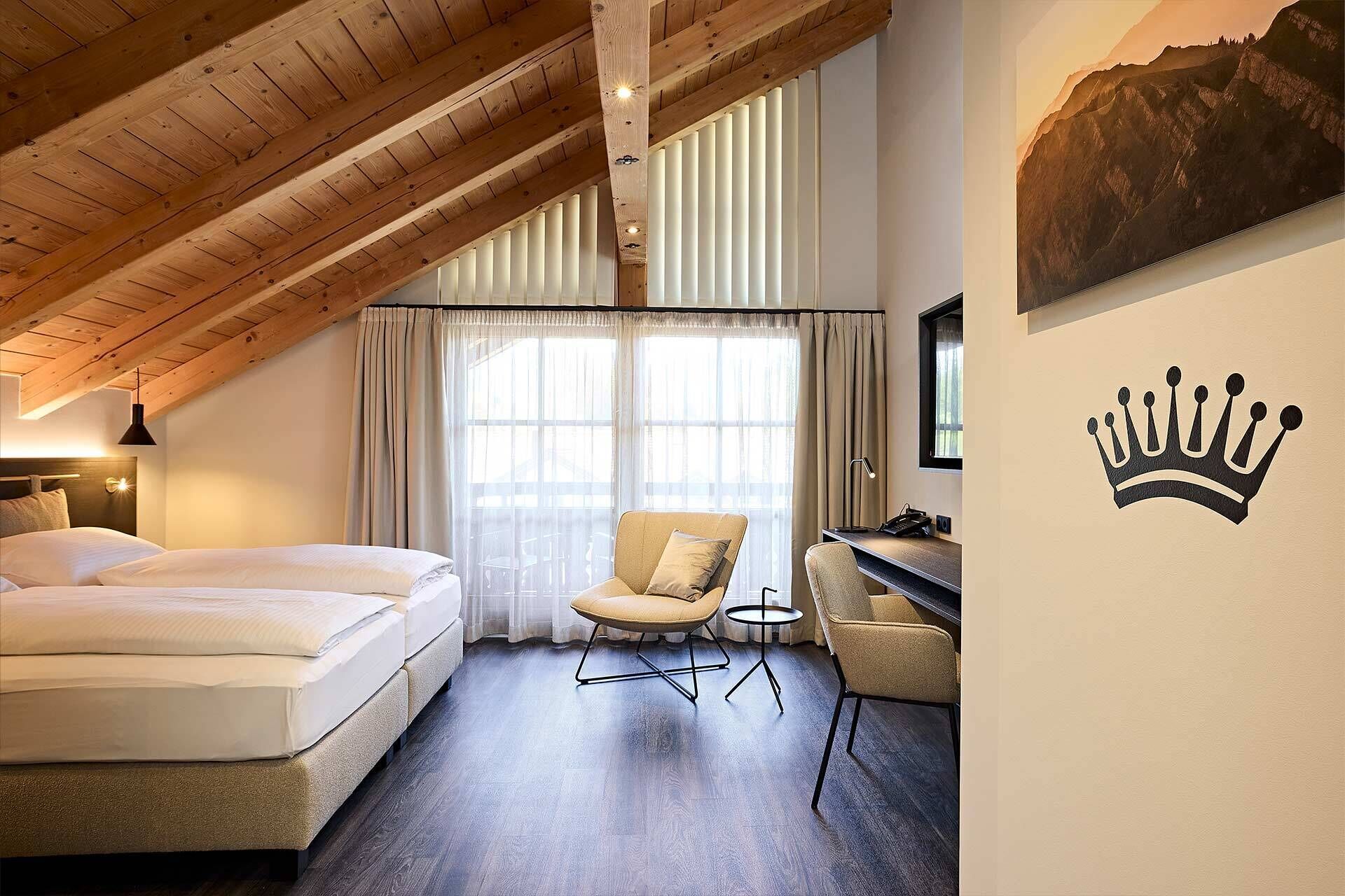 A bedroom with a wooden ceiling, a bed, a chair, a small table, and a wall with a crown design.