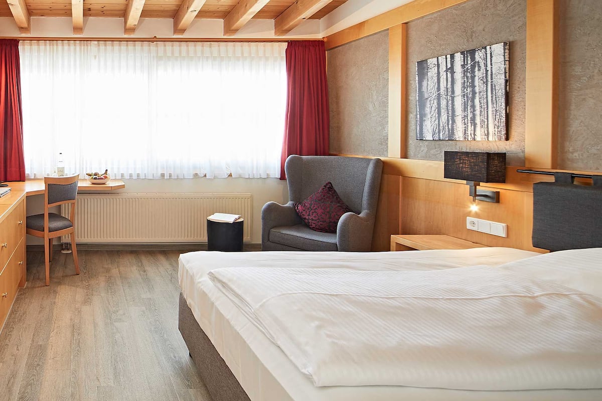 Hotellihuone, jossa on sänky, työpöytä tuolilla, pieni pöytä, seinään kiinnitetty lamppu ja ikkuna, jossa on verhot.