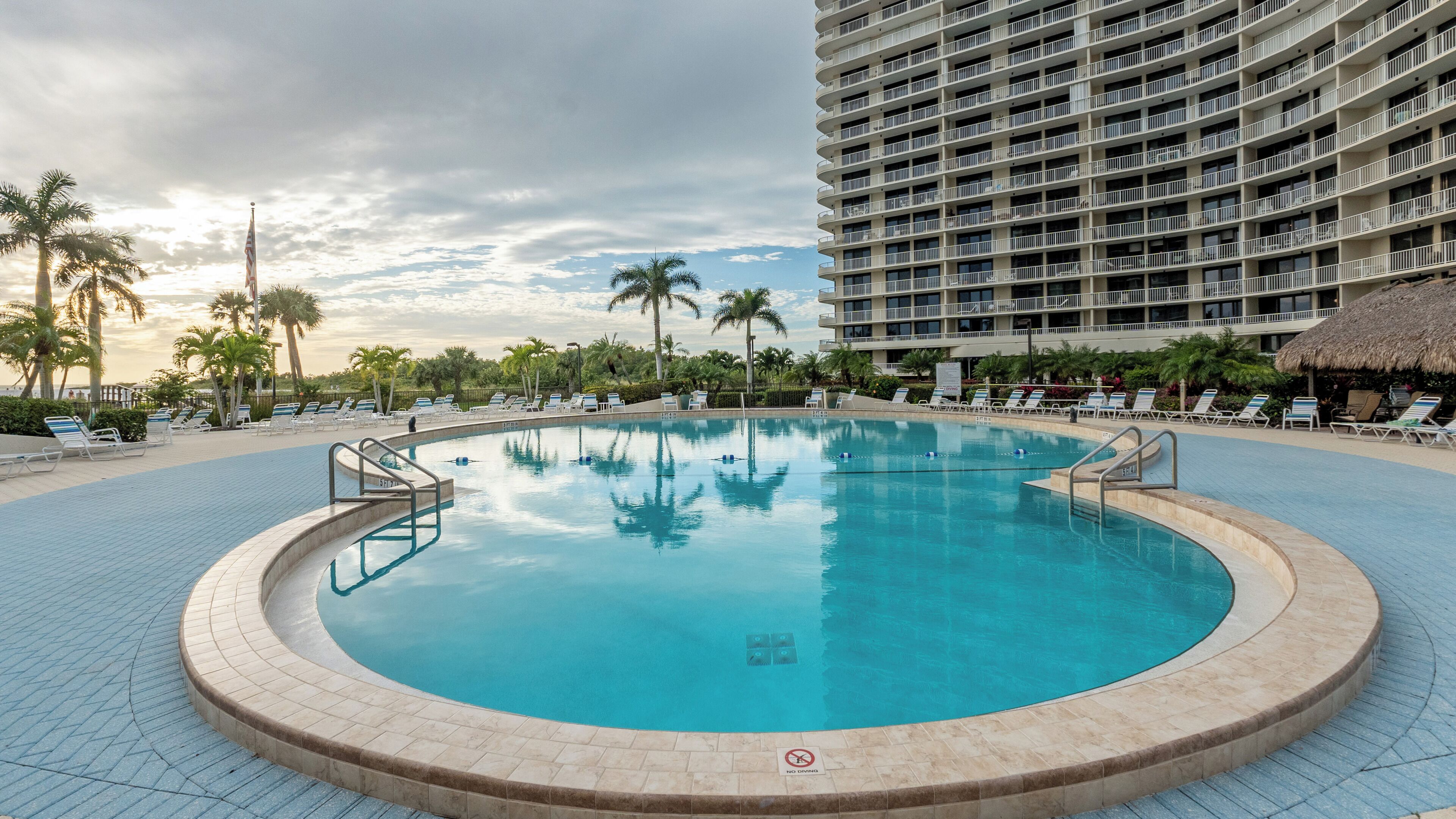 Uma grande piscina circular com placa indicando proibição de mergulho, rodeada por espreguiçadeiras e palmeiras, com um edifício alto ao fundo.