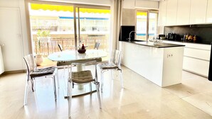 Una cocina moderna con una isla central, un comedor con sillas traslúcidas y vistas a un balcón con toldos amarillos.