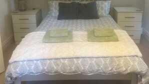 Un lit bien fait, recouvert d’une couvre-lit à motifs, avec deux oreillers et deux serviettes pliées. Il y a deux tables de chevet avec des lampes et un petit tiroir.