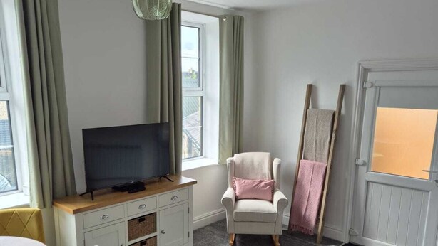 Un espace de vie compact comprenant un téléviseur posé sur un meuble en bois, un fauteuil blanc avec un coussin rose, un canapé gris et une petite table basse en bois.