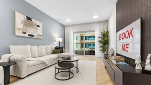 A modern living room with a sofa, two side tables, a coffee table, a TV, and a large window with a view of a building. - StayShort - Cozy Dubai Marina Stay with Private Balcony Sleeps 2 (Dubai Marina)