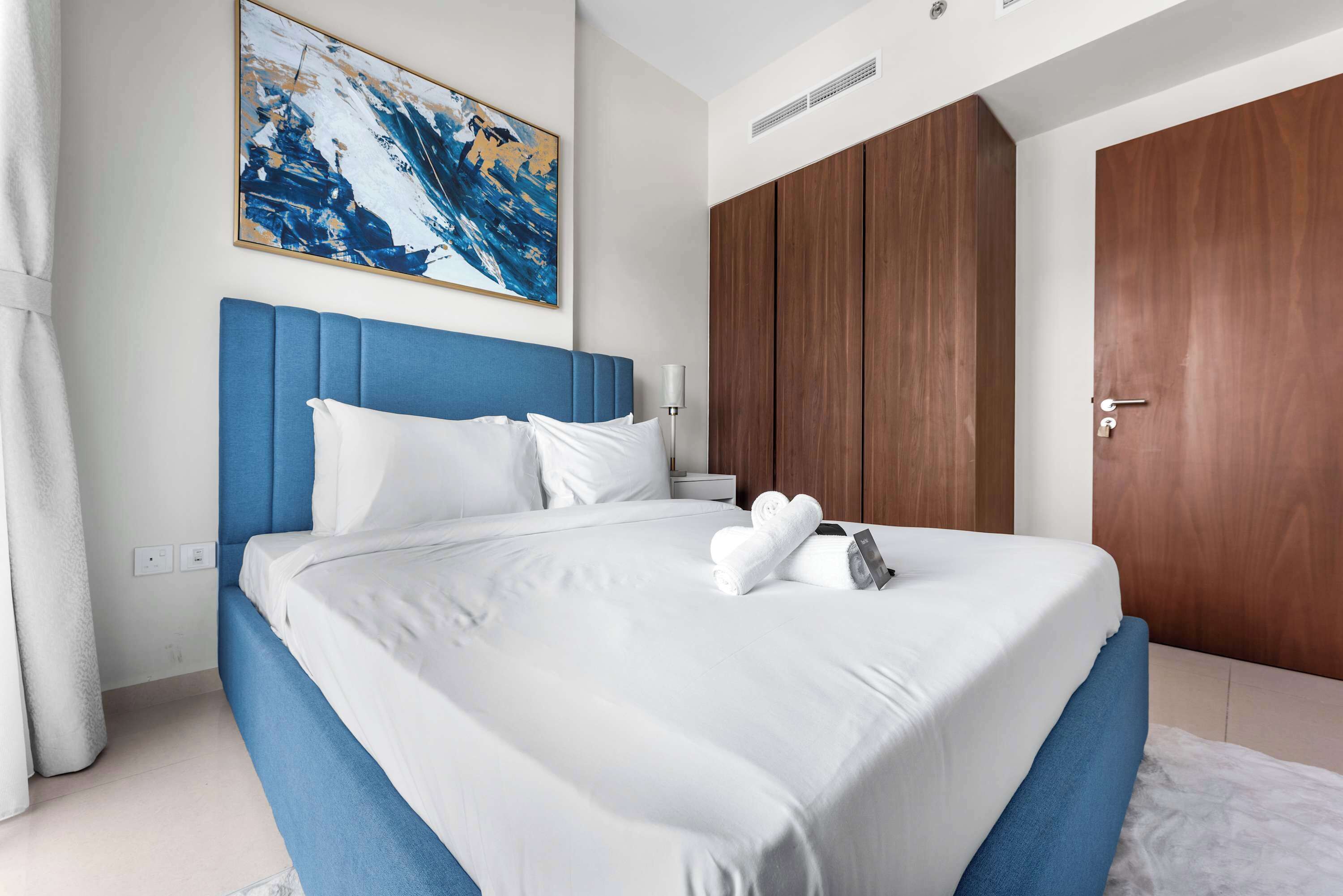 Una cama bien hecha con sábanas blancas y un cabecero azul, un armario de madera y un cuadro en la pared.