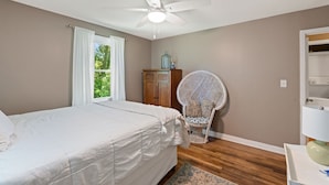 A bedroom with a bed, a ceiling fan, a window with curtains, a wooden cabinet, a wicker chair, and a small table with a lamp. - Home near DTWS, UNCAS, WFU, Hospitals, food/bev! (Winston-Salem)