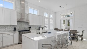 A modern kitchen with white cabinetry, a large island with bar stools, a stainless steel range hood, and a dining area with a chandelier. - Stunning New Main & Carriage House w/ Private Pool- Seas the Day of Seagrove (Santa Rosa Beach)
