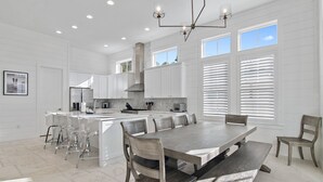 A spacious kitchen with white cabinetry, a large island with bar stools, and a dining area with a wooden table and chairs. - Stunning New Main & Carriage House w/ Private Pool- Seas the Day of Seagrove (Santa Rosa Beach)