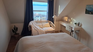A bedroom with a bed, a desk, a lamp, a potted plant, and a view of the sea.