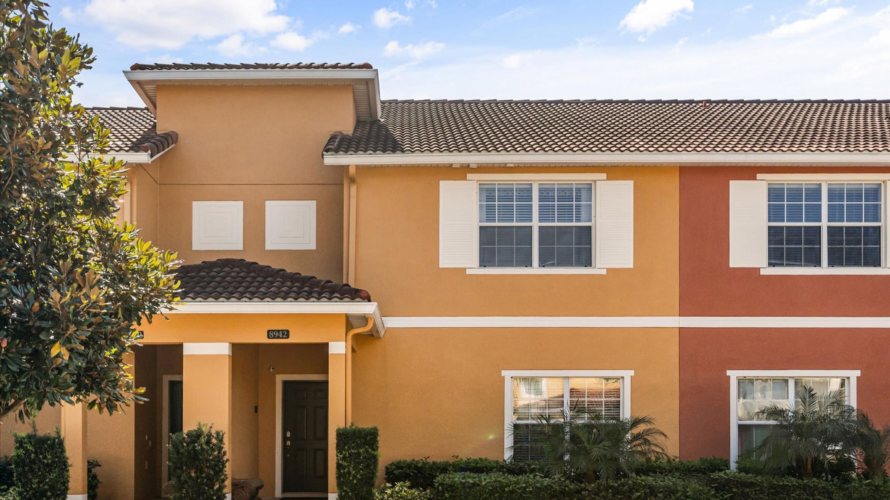 A two-story residential building with a beige and red facade, white shutters, and a tiled roof.