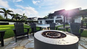 An outdoor patio with a stone floor, black Adirondack chairs, and a fire pit. - New 5 Bedroom Modern Villa on Golf Course (Fort Lauderdale)