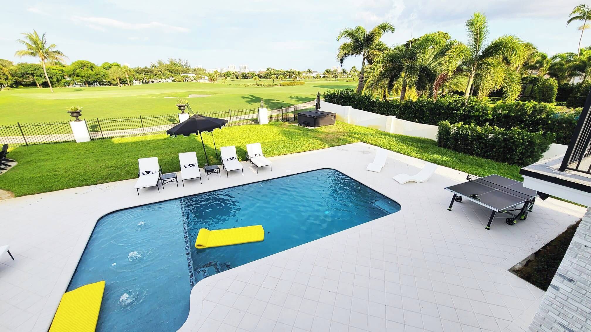 A swimming pool with sun loungers and a table, surrounded by a well-maintained lawn and palm trees.