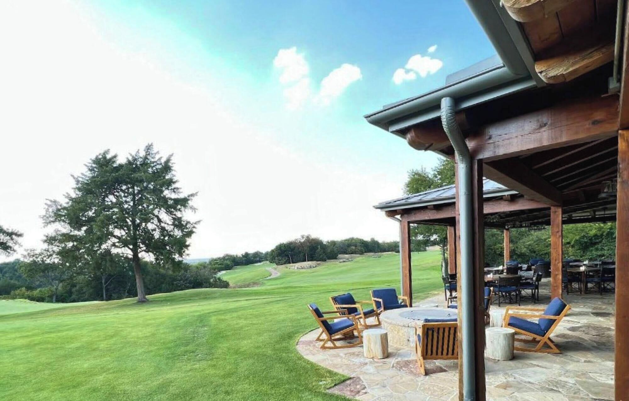 A covered outdoor seating area with stone flooring and wooden furniture, overlooking a well-maintained lawn and distant trees.