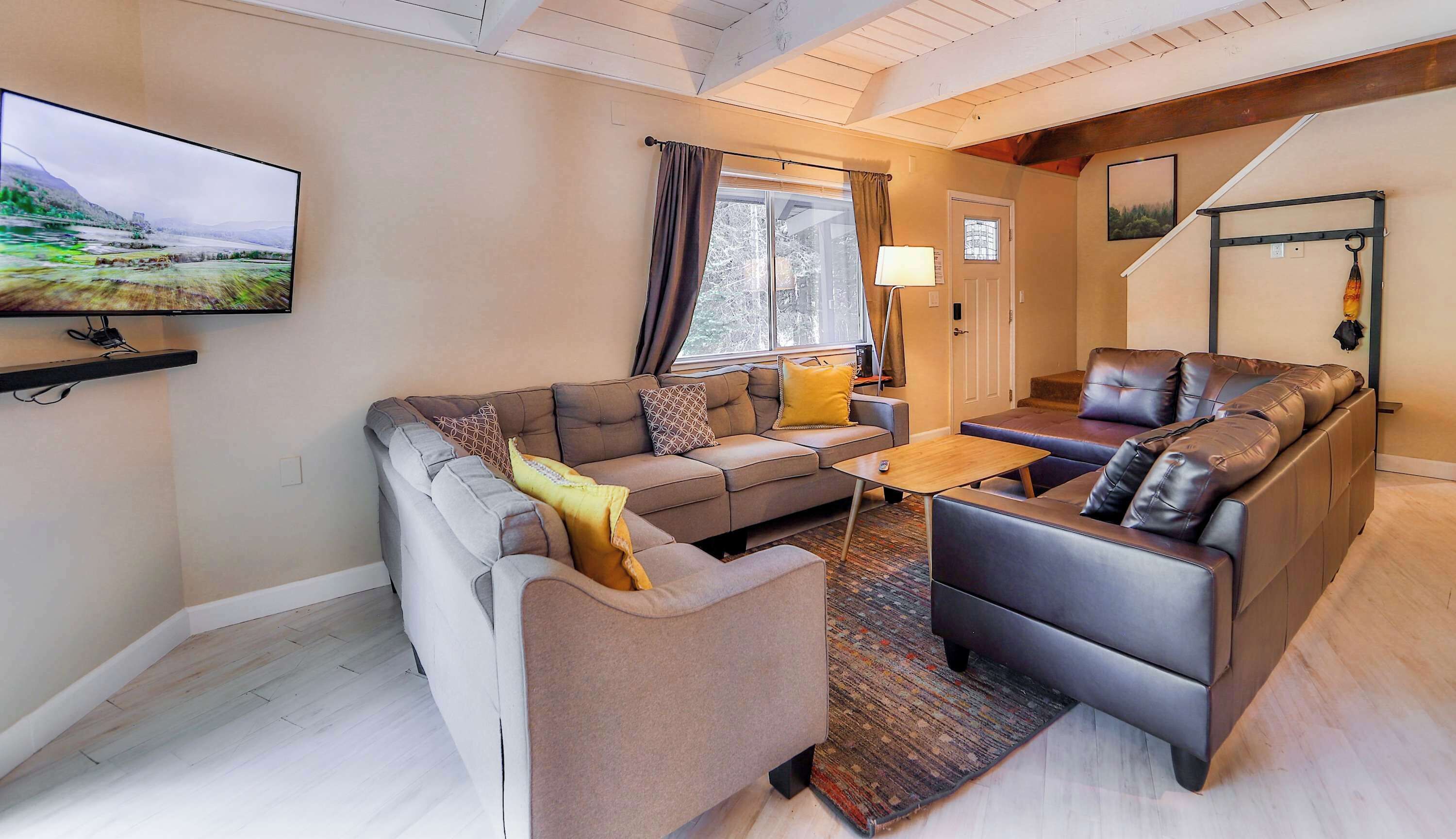 Een moderne woonkamer met een grijze bank, een bruine fauteuil en een salontafel. Er is een televisie aan de muur bevestigd, een raam met gordijnen en een trap op de achtergrond.
