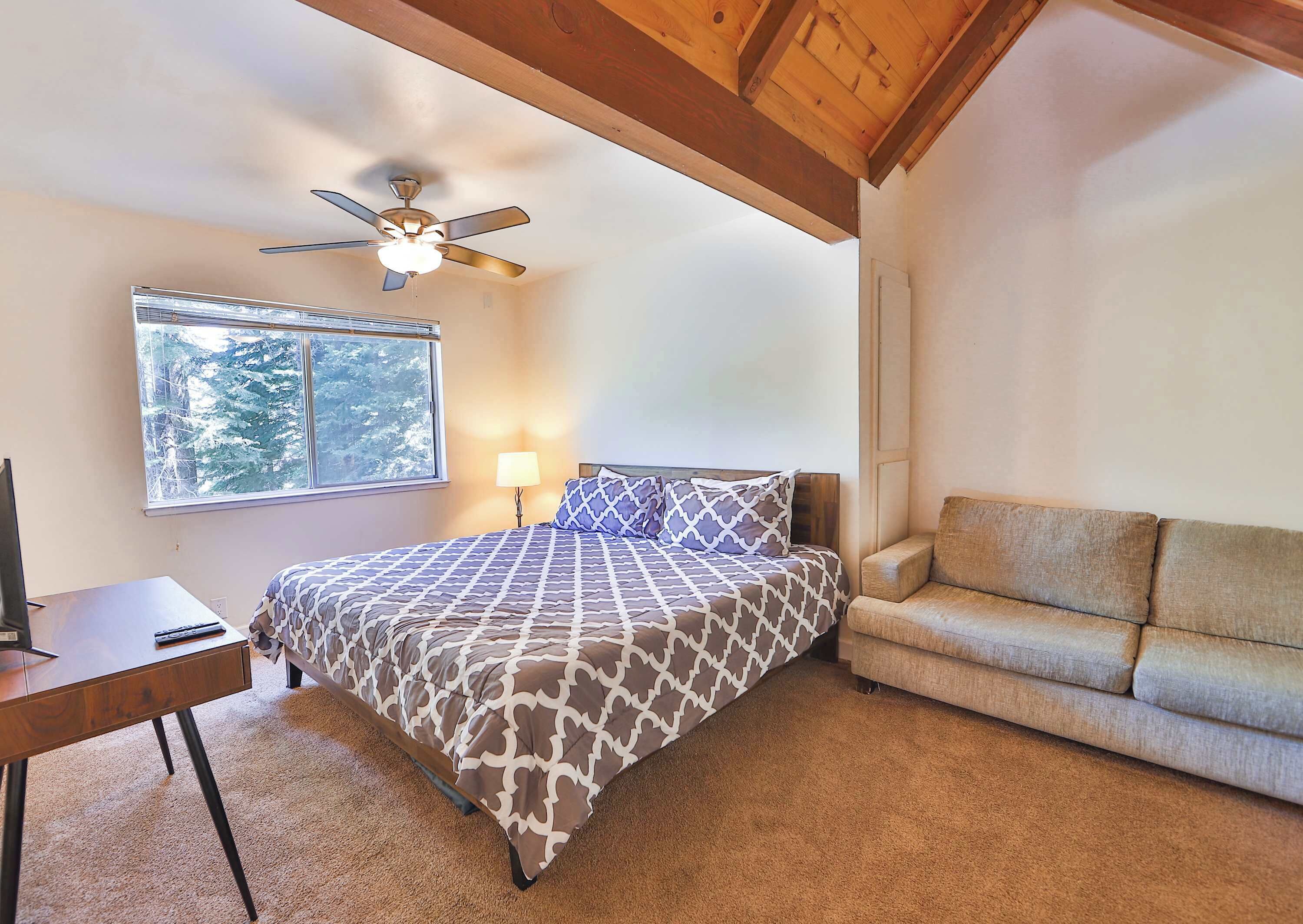 A bedroom with a bed, a desk, a chair, a ceiling fan, and a window with a view of trees.