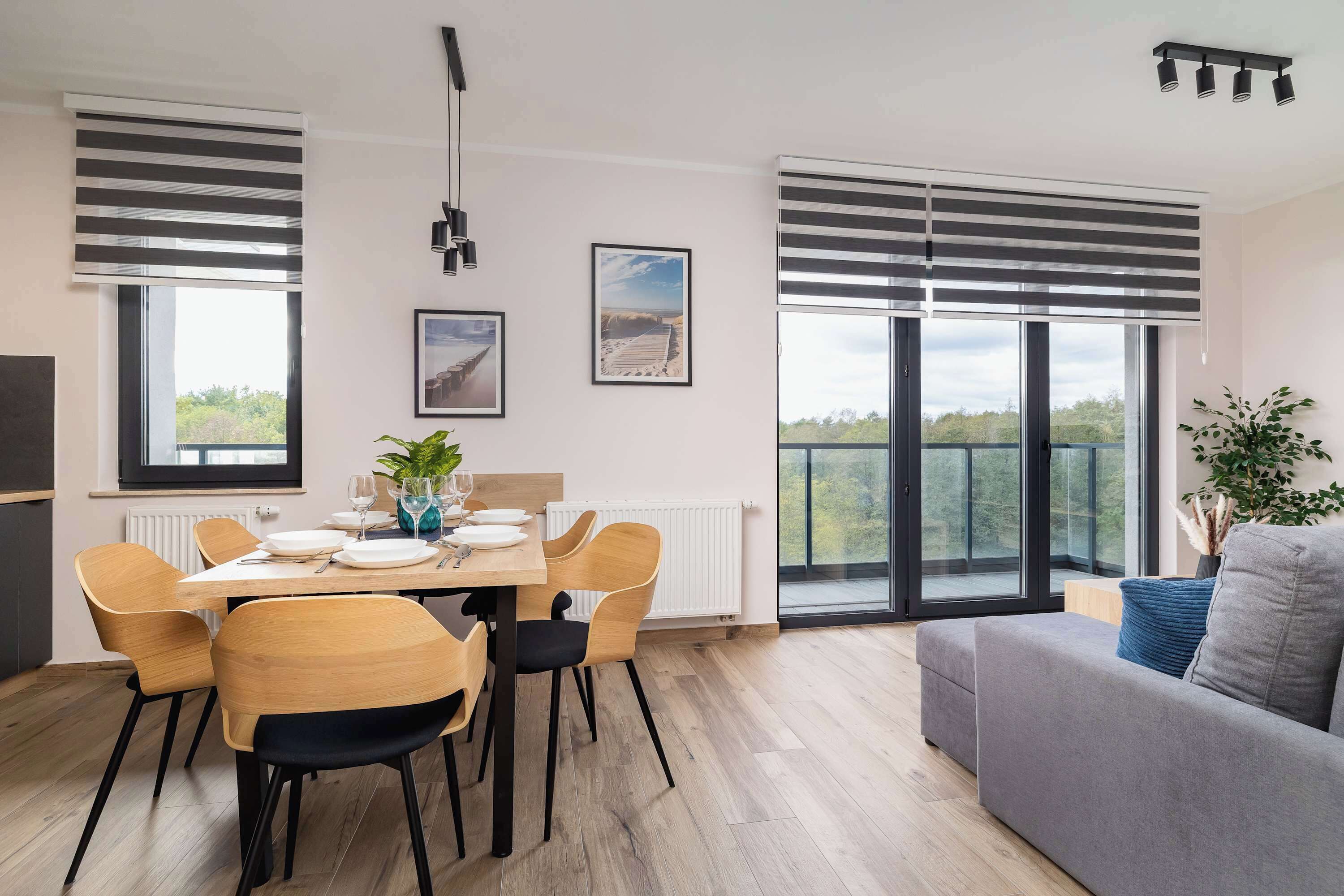 A modern dining area with a wooden table and chairs, large windows with blinds, and a grey sofa.