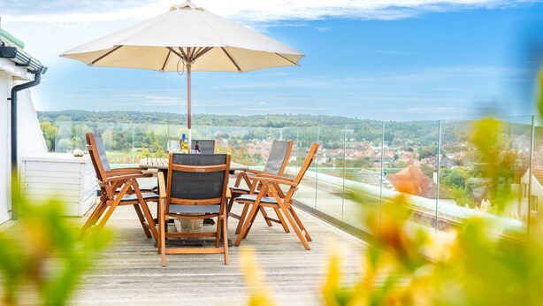 A wooden deck with a table and chairs, an umbrella, and a glass railing overlooking a scenic landscape. - Greentiles, Sheringham, Norfolk (Sheringham)
