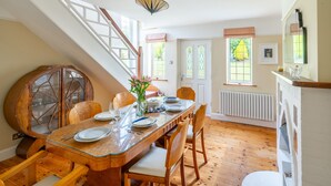 A dining area with a wooden table set for four, surrounded by chairs, a vase with flowers, and a staircase leading to an upper level. - Greentiles, Sheringham, Norfolk (Sheringham)