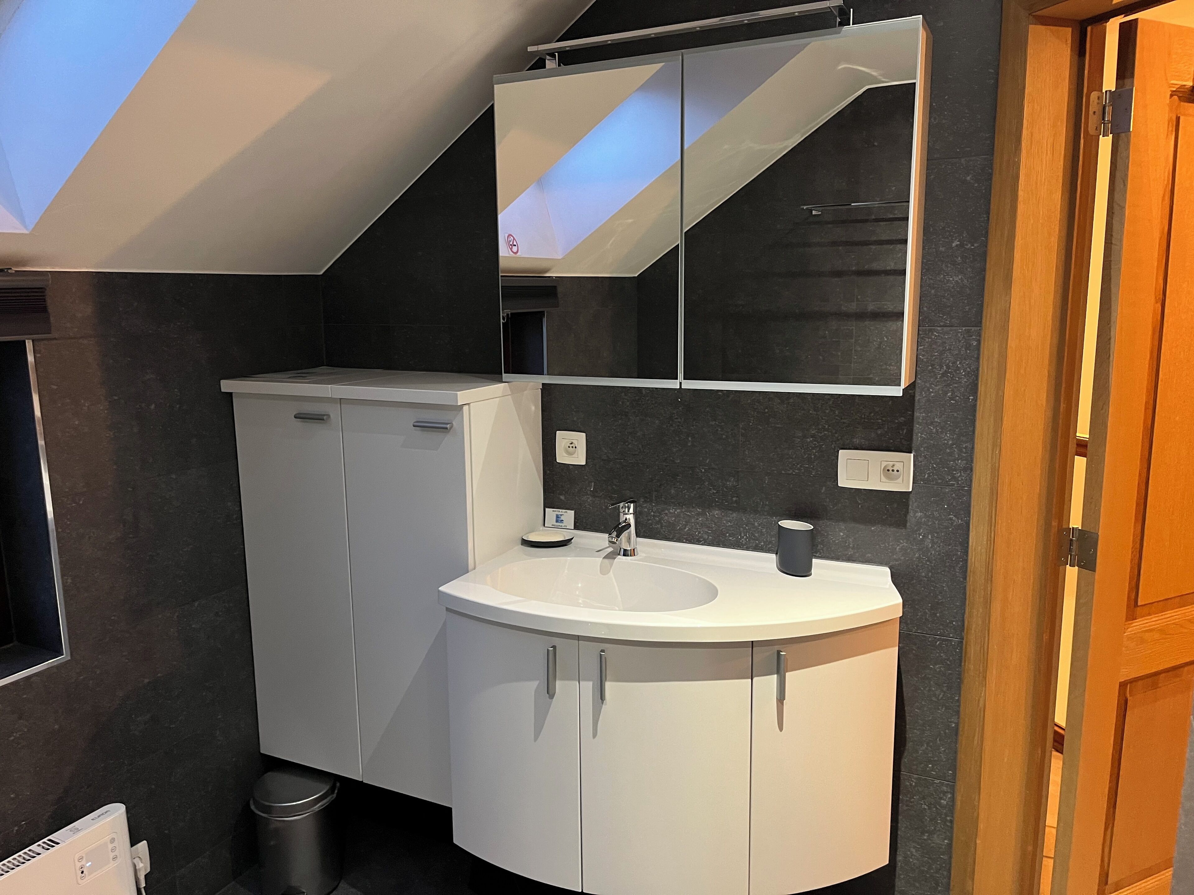 Une salle de bain moderne avec un lavabo blanc, un miroir et un mur recouvert de carreaux noirs.