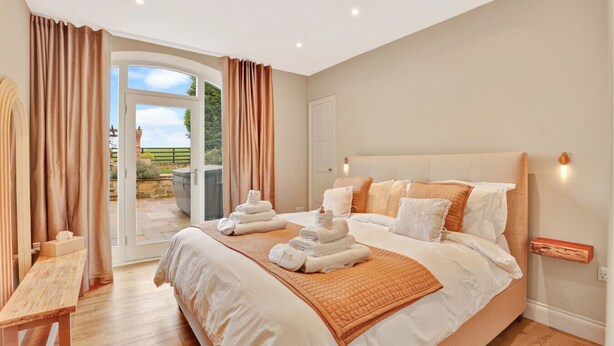 A spacious bedroom with a large bed, a wooden bench, and a view of the outdoors through a glass door. - The Retreat at Glenridge House - North East Escapes (Wooler)