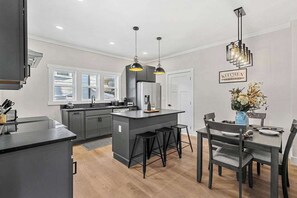 A modern kitchen with grey cabinets, a central island with stools, pendant lighting, and a dining area with a table and chairs.