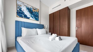 A neatly made bed with white linens and blue headboard, a wooden wardrobe, and a painting on the wall. - Stayshort - Business Bay for 4 People With a Fabulous Pool (Dubai)
