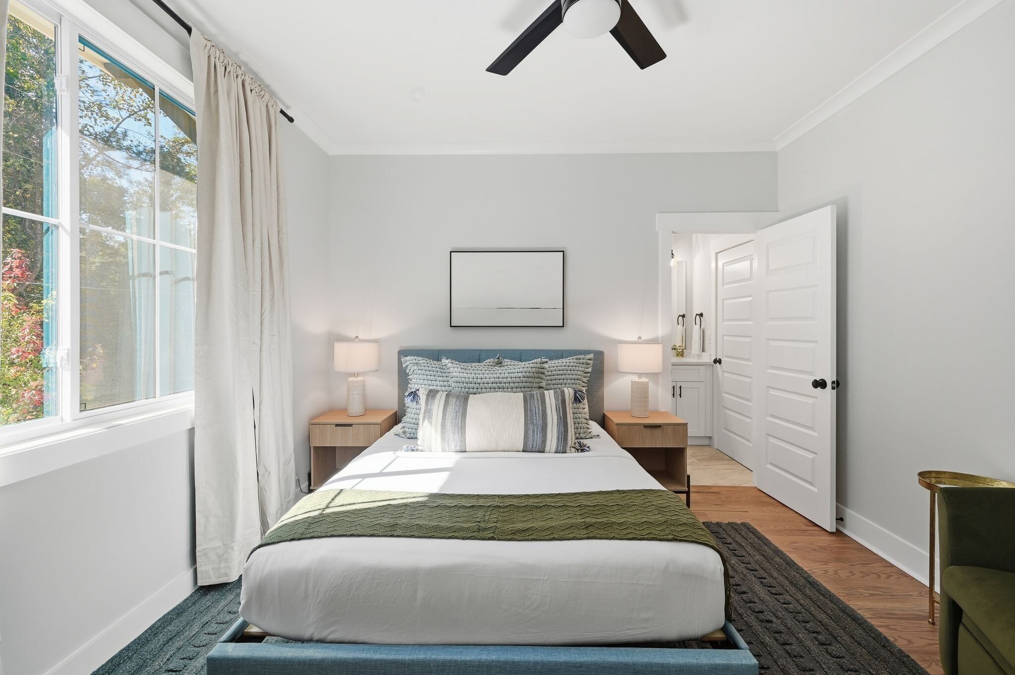 A bedroom with a bed, bedside tables, a ceiling fan, a window with curtains, a door, and a wall-mounted picture.