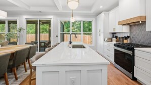 A modern kitchen with a large island, white cabinetry, and a view of the backyard through large windows. - New! Modern Luxurious Woodland Retreat 5 Bedroom Home (Scottdale)