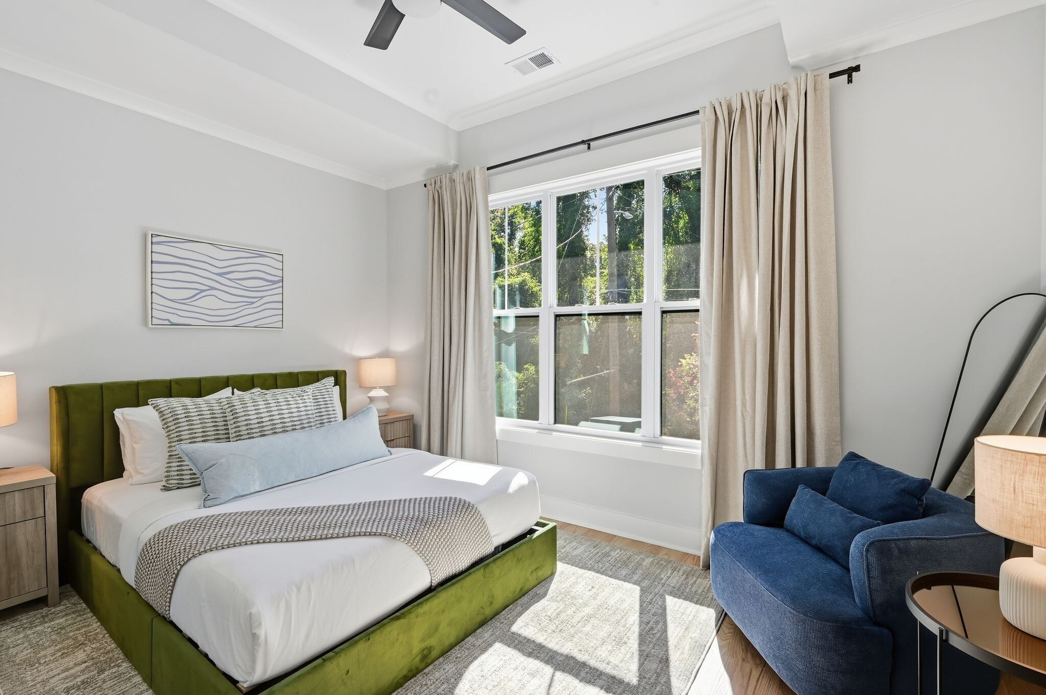 A modern bedroom with a green upholstered bed, a blue sofa, and a large window with curtains.