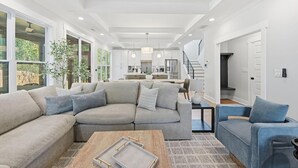 A spacious living area with a large L-shaped sofa, a coffee table, and a blue armchair. A kitchen is visible in the background with modern appliances and a dining area. - New! Modern Luxurious Woodland Retreat 5 Bedroom Home (Scottdale)