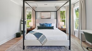 A four-poster bed with a view of greenery outside, a bedside table with a lamp, and a small ottoman. - New! Modern Luxurious Woodland Retreat 5 Bedroom Home (Scottdale)