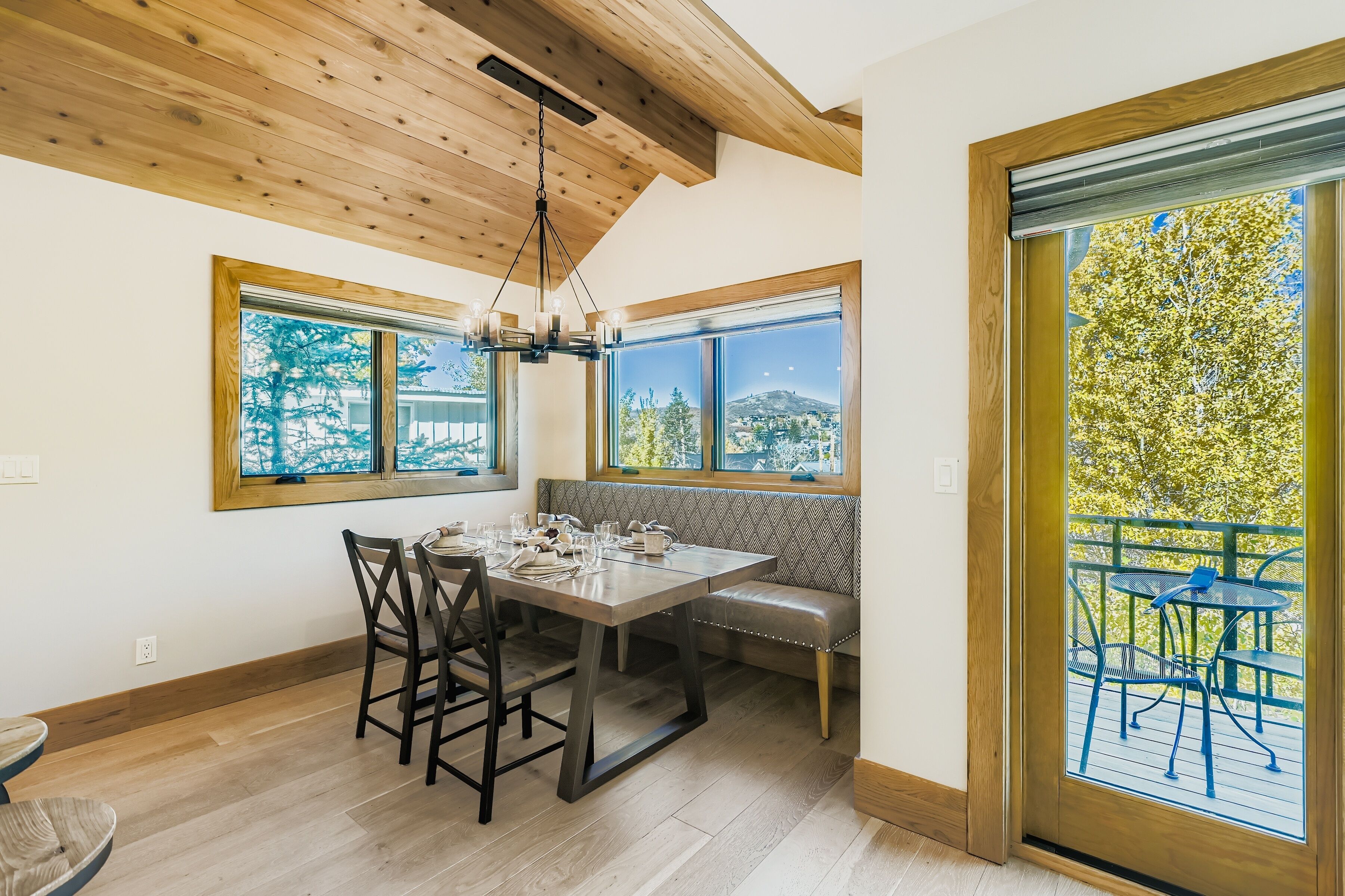 Un comedor con techo de madera, ventanas amplias con vista a árboles y una montaña distante, un banco y una mesa preparada para cuatro.