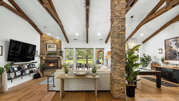 Un salon spacieux doté d’une cheminée, de poutres en bois, de piliers en pierre et d’une grande fenêtre donnant sur un jardin.