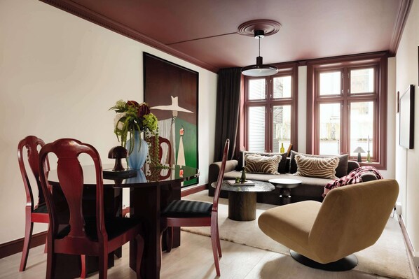 A dining area with a table, chairs, and a vase of flowers. There is a sofa and a lamp in the background.